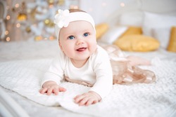 Baby girl wearing cute dress and headband, lies on a white cover in festively decorated room. With surprise watches in the camera, on a background a set of bright fires, soft focus