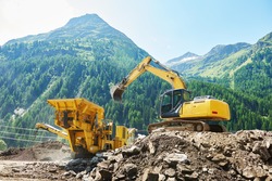 excavator loads the ground in the stone crusher machine during earthmoving works outdoors at mountains construction site