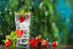 refreshing carbonated drink with many strawberries and ice cubes, against a background of greenery and falling drops, summer concept, copy space