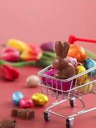 Close up of Easter rabbit chocolate in a mini shopping cart with colorful Easter egg, and festive decoration atmosphere. Colorful tulips flower with pink background