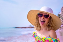 Happy little girl makes a funny grimace and shows her tongue. Adorable girl in a big straw hat, sun glasses and in a swimsuit relaxing on the beach near sea. Cute child on vacation looks at the sea.