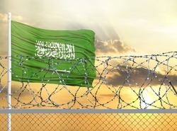 Flagpole with the flag of Saudi Arabia against the sky and behind a fence with barbed wire. The concept of protecting the borders of territories.