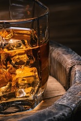 Glass of amber-colored whiskey stands on barrel of whiskey with soft focus close-up. Brandy with ice alcoholic drink with aroma of cognac.