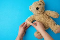 two hands holding a teddy bear and gluing an adhesive plaster on a paw, blue background, top view