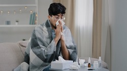 Arab bearded sick sad man with glasses sits on couch at home suffers from runny nose flu disease coronavirus pandemic covid epidemic sneezes. Unwell guy feeling bad fever virus illness symptoms indoor