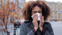Young beautiful girl teenager millennial African American woman with curly hair stands in city on street suffers from seasonal allergy rhinitis runny nose virus illness sneezes wipes with paper napkin