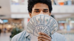 Happy wealthy successful smiling african american business man looking at camera holding fan of money hiding behind dollar banknotes showing financial profit winning salary savings loan credit cash