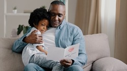 Mature african american man with cute kid girl hugging sitting in room on sofa father reads greeting card loving daughter made gift with own hands for birthday or father's day dad happy cuddling baby