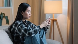 Side view of sad bored Asian woman girl sitting on sofa at home with telephone serious unhappy lady scrolling browsing in smartphone and using mobile phone app watching social media news feed typing
