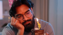 Headshot arabian alone single sad upset lonely indian bearded man in festive birthday pink hat and glasses sitting at home at night feel frustrated despair disappointed sadness holds cake with candle