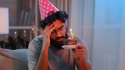 Portrait Arabian sad upset lonely Indian bearded man celebrate birthday alone without friends feel sadness headache failure male in festive pink hat and glasses at home at night with cake with candle