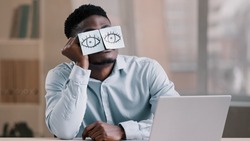 Sleepy funny african american office manager business man sitting at home workplace with sticky notes on eyes comic young tired worker has sleeping problem napping at desk wears glasses with stickers