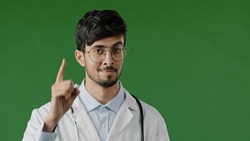 Arabic doctor man smiling hispanic male medical worker raises index finger warns you to be attentive disbelief looks at camera express ironic emotion stands over green background shows caution