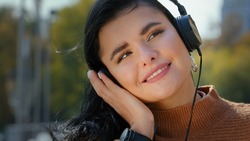 Portrait young happy woman standing outdoors in headphones listening favorite music using smartphone carefree hispanic girl smiling dancing to pleasant song melody audio sound female enjoying life