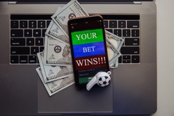 Smartphone with online betting application, dollar bills and soccer ball on a laptop. Gambling concept