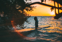 Silhouette of the loving couple standing barefoot on the ocean pier, hugging and kissing each other during a stunning orange sunset over the seascape, red lens flare, branches of trees around