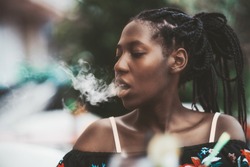 Portrait of a dazzling African girl with braids sitting in a street cafe and exhaling smoke from the hookah; young black female outdoors in a bar plays with the vapor from an electronic cigarette