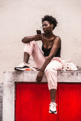 A vertical view of a charming young African female in eyeglasses, white trousers, and a black top tank is sitting on the top of red gates and using her smartphone; black girl using cellphone outdoors