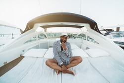 A cheerful wealthy bearded mature black guy in an elegant striped black and white costume with shorts and hat is sitting outdoors on the deck of a luxury bright speed yacht moored on a berth