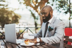 A cheerful handsome bald black businessman in eyeglasses and neat beard is having a coffee break in an outdoor restaurant, pouring himself some tea while reading a chat message on his laptop in front