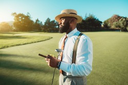 A portrait of a handsome dapper bearded black man in an elegant outfit: white shirt, trousers with suspenders, tie, and hat, is standing on the golf field and holding the club, cigar, and ball in hand