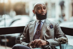 Portrait of a dapper mature hairless black man entrepreneur with a nice beard, in a custom elegant grey costume, sitting in an outdoor cafe on a rainy day and pensively looking into the distance
