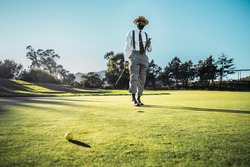 A fashionable good-looking mature bearded black guy in pants with suspenders and a straw hat, with a cigar and a golf club, is standing on the green grass of the lawn on a golf field on a warm evening