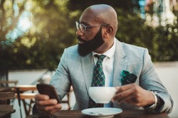 A portrait of a handsome dapper bald mature black man entrepreneur with a well-groomed beard and in a formal suit with a necktie, sitting in a street restaurant and drinking tea during a coffee break