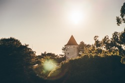 A landscape with a selective focus on a small tower of a castle or some antique building surrounded by trees and greenery and backlit by the evening sun making a lens flare and a chroma hoop