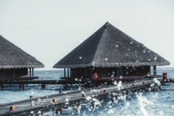 View of typical luxury triangle cottage houses on a Maldives resort with triangle canopy roofs and a pedestrian pier or a bridge with water splashes in a defocused foreground on a bright warm day