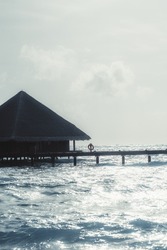A vertical shot of a seascape with a canopy bungalow of a luxurious Maldives resort with a triangle roof and a wooden pier with a lifebuoy on it on a warm bright day with highlights on the waves