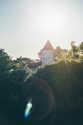 A vertical contrast shot with a tower of an antique building or a castle with a conical triangle roof backlit by bright evening sun casting lens flares, with forest greenery in the foreground