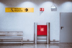 An interior of an airport terminal or a subway train station with a red fire hydrant cabinet, a yellow pointer to phones, a service entrance door, and a chrome bench in front of a white tiled wall