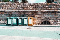 On the boardwalk of Cascais, a handful of eco-points are positioned against a sedimentary calcareous brownstone wall. They are located near an underground passageway with iron grates.