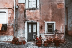 In Lisbon, a weathered house with cracked walls and peeling paint stands on a black or dirty cobblestone floor. Adorned with clay pot flowers, it exudes a sense of aging beauty and rustic charm