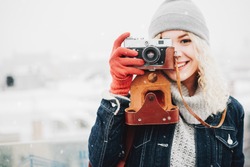 Young blond curly smiling female in warm clothes with retro film camera shooting a photo on the background of winter city, snow