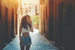 Beautiful curly woman talking with her boyfriend while standing on dark narrow street in summer day, stylish hipster brunette girl is using smartphone for calling her family during waiting her friend