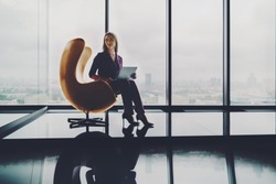 Beautiful woman entrepreneur is sitting alone with laptop on modern yellow curved armchair in office interior with reflections on high floor of skyscraper with copy space place for logo or your text