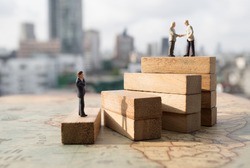 Miniature people: Successive business concept. Businessman thinking on first step of wood stair and two businessmen shaking hands on top step with modern city background.