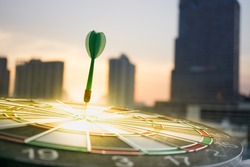Green dart arrow hitting in the target center of dartboard with modern city and sunset background. Target business, achieve and victory concept .