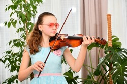 A girl in sunglasses plays the violin at home among green plants.