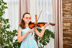 A girl in sunglasses plays the violin at home among green plants.