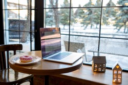 Workplace with a laptop. A table with a large window and a donut. Co-working. Workspace in a cozy coffee shop. It is snowing outside the window. Work on a laptop in winter
