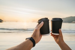 Two disposable paper cups in the hands of a man and a woman against the backdrop of a beautiful sunset and sea landscape. The concept of love, relationship or Valentine's Day celebration.