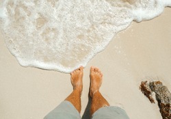 Male feet barefoot stand on the sand of the beach with sea foam. Tropical travel and vacation concept.