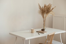 Aesthetic minimal office workspace interior design. Mug, notebook, pampas grass floral bouquet on white table against white wall. Girl, woman boss work at home business concept.