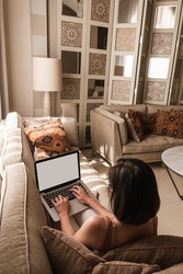 Young beautiful woman work on laptop computer with blank screen. Eastern traditional home interior design concept. Oriental style living room with marble table, sofa, pillows, shutter, decorations