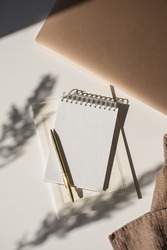 Spiral flip notebook with blank copy space, pen, laptop computer in flowers sunlight shadow on white table. Aesthetic bohemian minimalist workspace. Artist, writer template