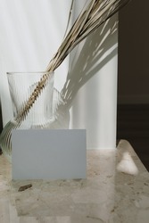 Blank paper card sheet with empty copy space. Dried palm leaf in stylish vase on marble table. Shadows on the wall. Silhouette in sun light. Branding business invitation card with copyspace
