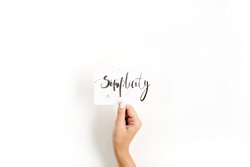 Minimal pale composition with girl's hand holding card with word Simplicity written in calligraphic style on paper on white background. Flat lay, top view